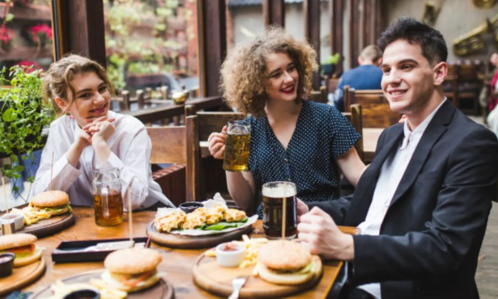 Three people sit at an outdoor cafe table with burgers and fries, smiling and chatting