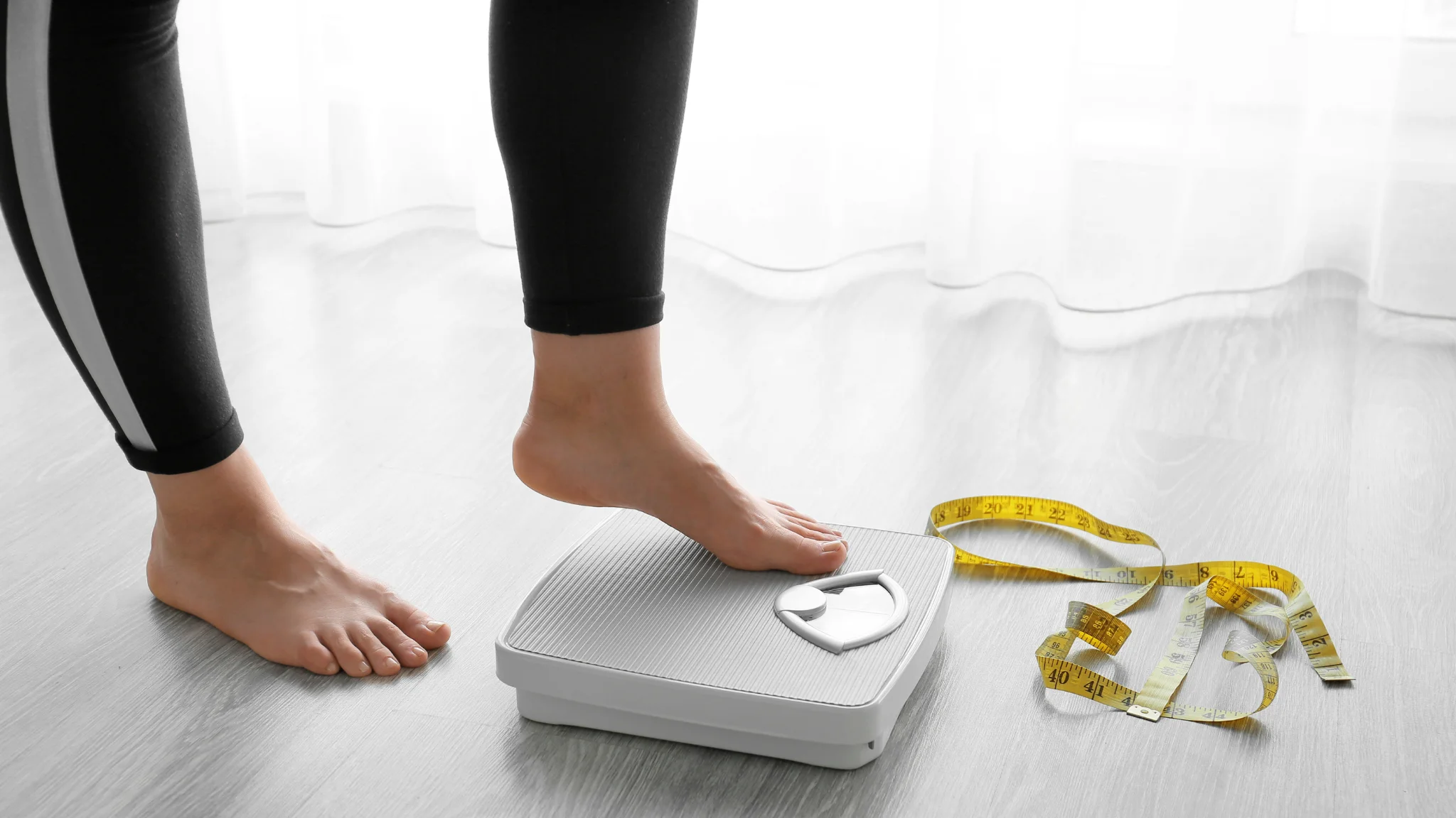 A woman stands on a scale, holding a tape measure, assessing her weight and measurements.