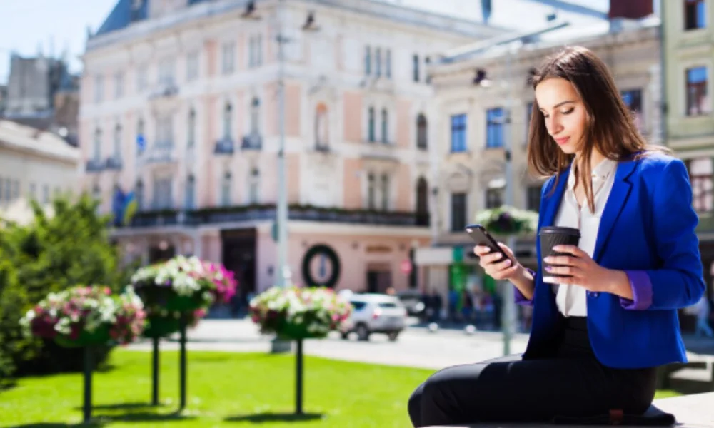A woman in a blue blazer sits on a ledge in a sunlit city square