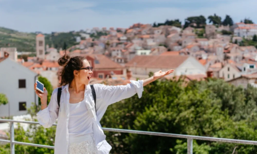 A woman stands excitedly on a terrace, holding a phone and gesturing towards a scenic view of a sunlit