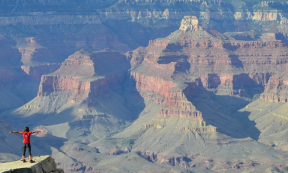 A person in a red shirt stands on a cliff at the edge of the Grand Canyon, arms outstretched-places to visit in arizona