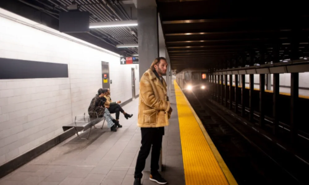 A man in a tan coat stands on a subway platform-planning a trip to new york