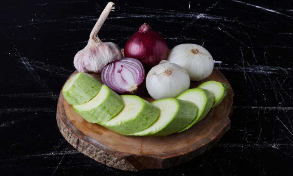 Sliced zucchini, whole and halved onions, and a garlic bulb on a wooden board-potato soup recipe