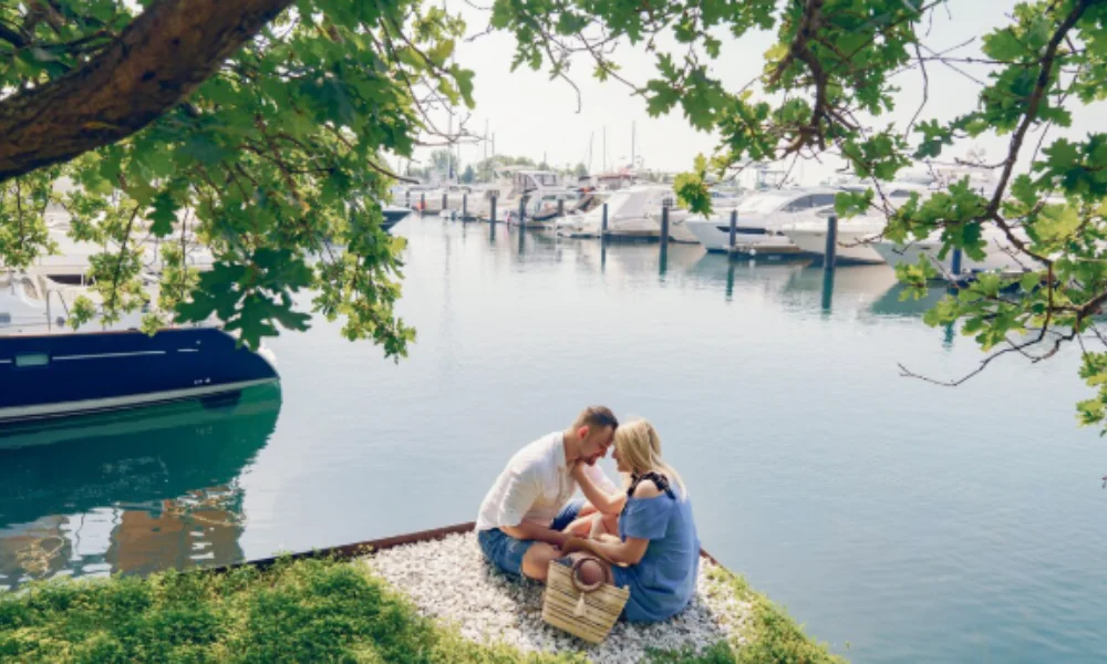 A couple sits close on a small patch of grass by calm marina waters-places to visit in italy