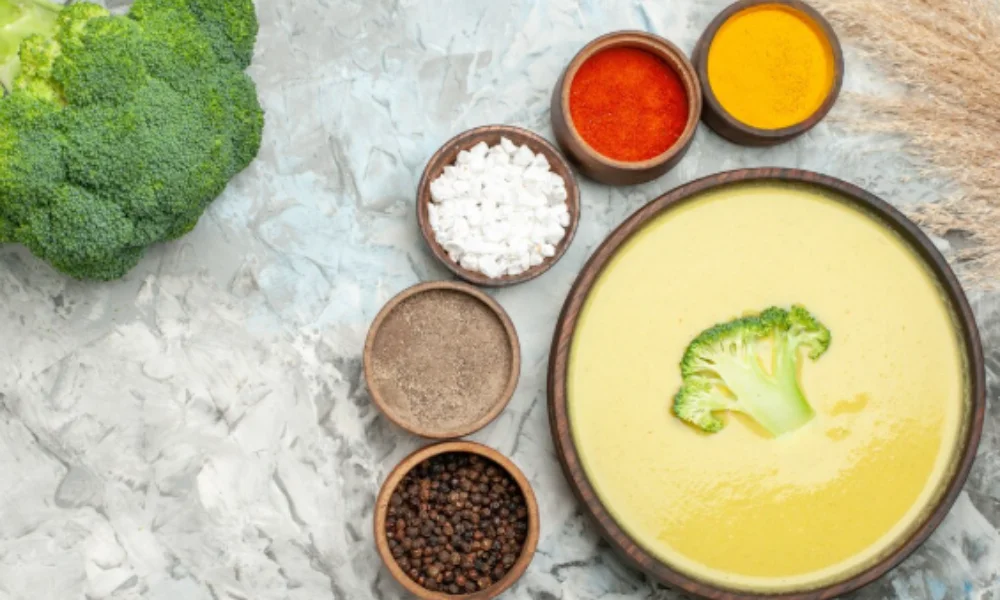 A bowl of creamy yellow soup garnished with a broccoli floret is surrounded by spices-potato soup recipe