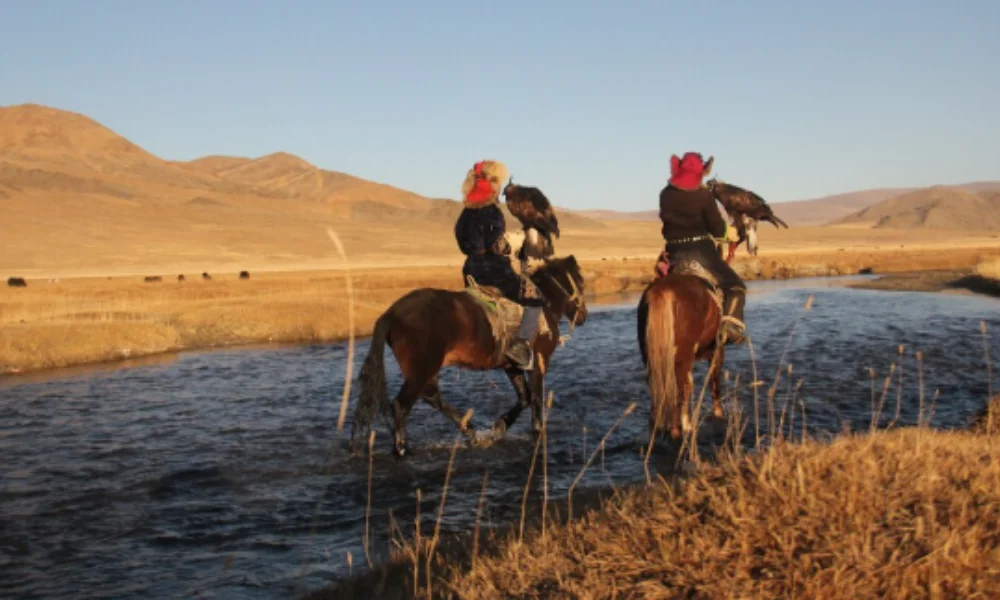 Two riders in traditional attire hold eagles on horseback by a tranquil stream in a vast