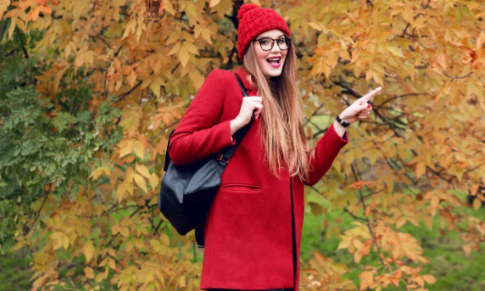 A smiling woman in a red coat and beanie points playfully in an autumn park