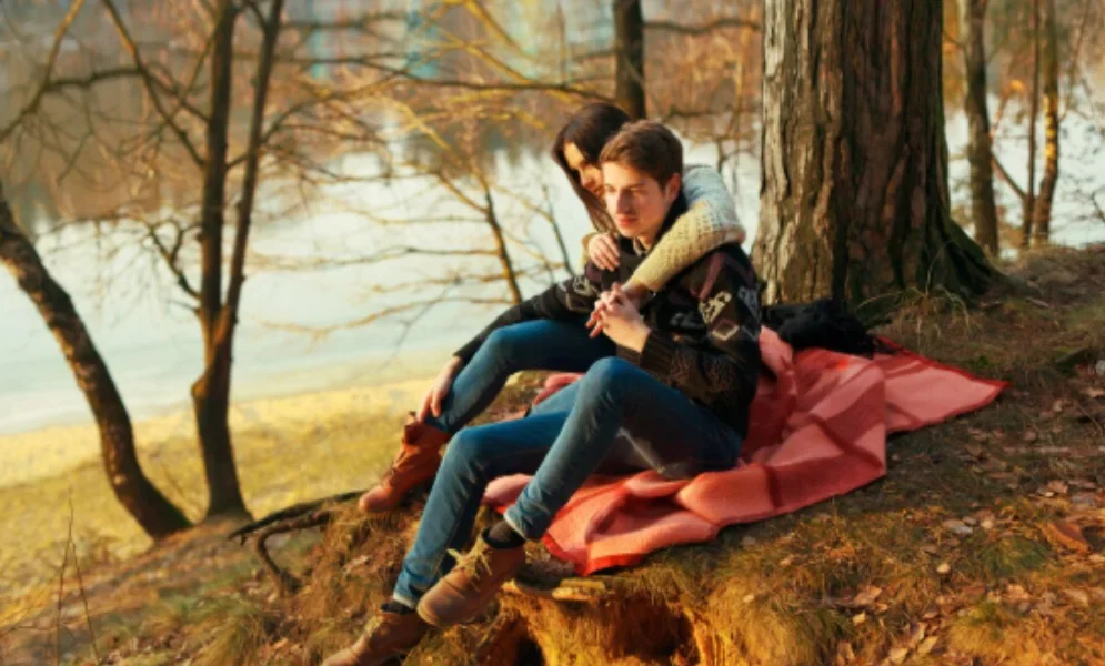 A couple sits on a red blanket in a peaceful forest by a lake