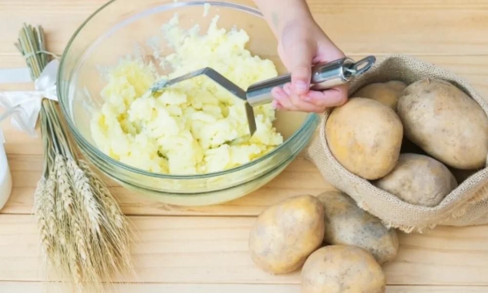 A hand mashes potatoes in a glass bowl on a wooden table