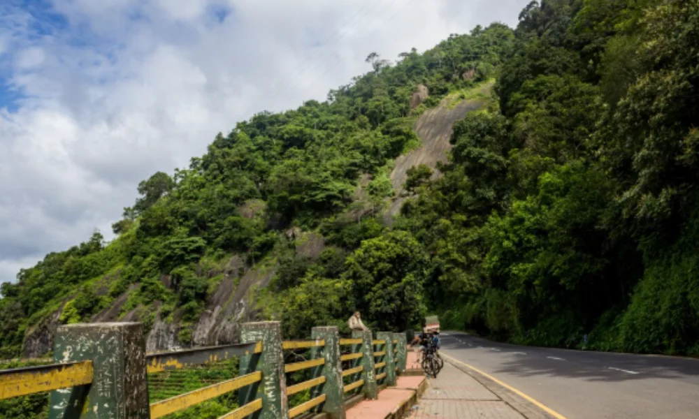 A winding road curves alongside lush, green hills under a partly cloudy sky