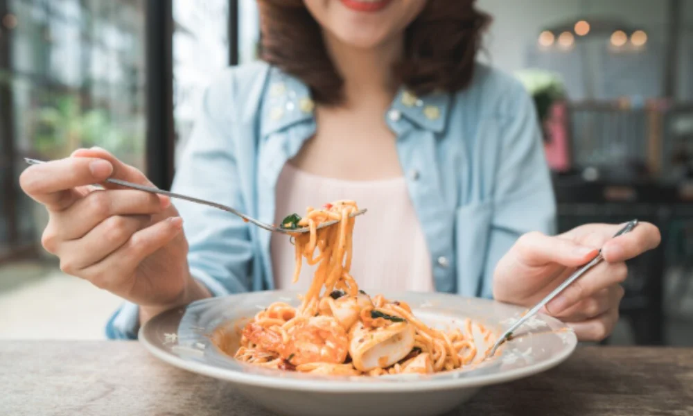 A person in a blue shirt, smiling, is using a fork and spoon to eat spaghetti with seafood in a bright