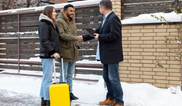 A couple with a yellow suitcase greets a man with a tablet on a snowy sidewalk-Cheap Destinations to Travel in December