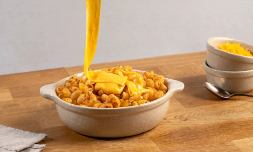Creamy cheese sauce is being poured over a bowl of cooked pasta shells on a wooden table