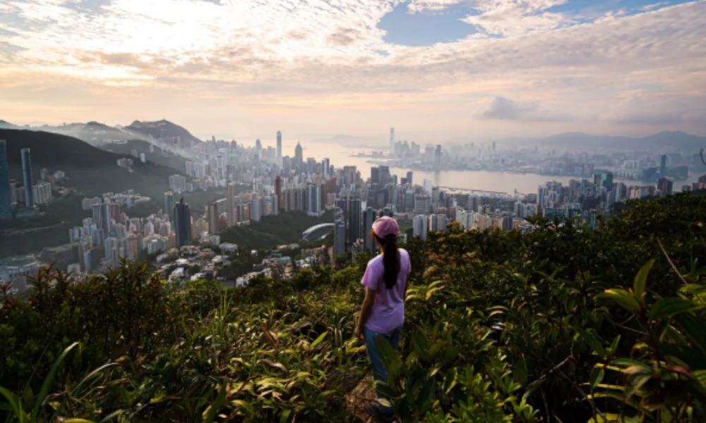 A woman in a pink hat and shirt stands on a lush hill overlooking a sprawling cityscape
