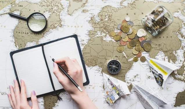 A hand writes in an open notebook on a world map. Nearby are coins-travelling around the world