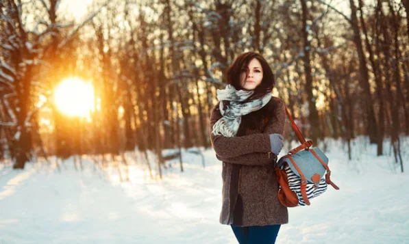 A woman stands in a snowy forest at sunset, wearing a coat and scarf, holding a bag
