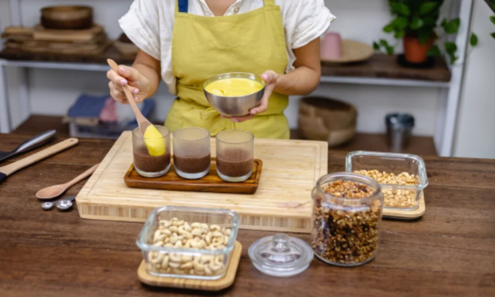 A person in a yellow apron adds yellow mousse to glasses with chocolate layers on a wooden tray