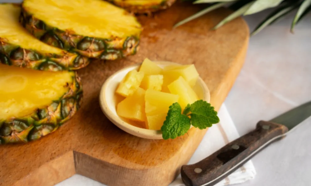 Slices of fresh pineapple on a wooden cutting board with a bowl of chopped pineapple garnished with mint leaves