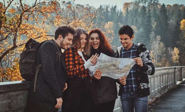 A group of four friends joyfully examines a map on a bridge surrounded by colorful autumn trees and a forest