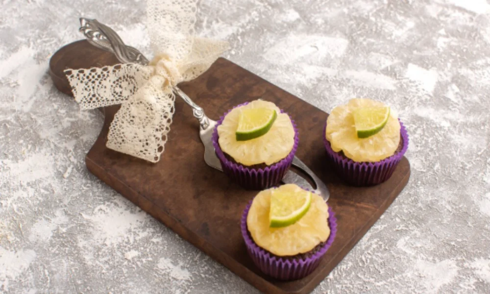 Three pineapple-topped cupcakes with lime slices on purple wrappers sit on a wooden board adorned with lace ribbon