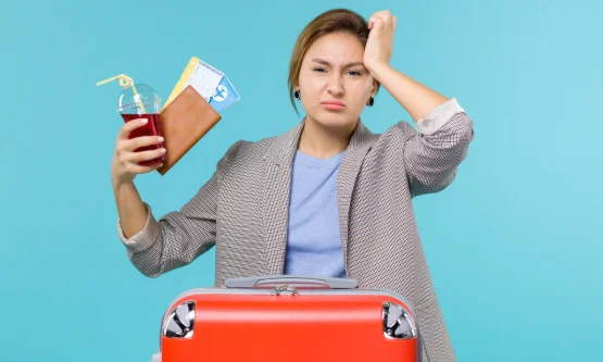 A frustrated woman stands beside an orange suitcase, holding a drink 
