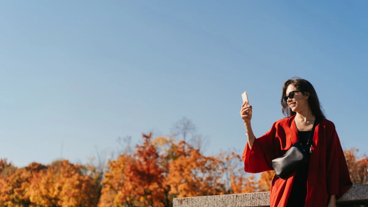 A woman in a red coat takes a selfie with her phone on a sunny autumn day. She stands near a ledge