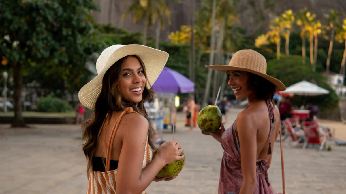 Two smiling women in sundresses and wide-brimmed hats drink from coconuts on a tropical beach path