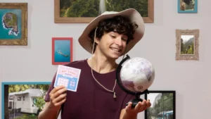 A young man with a wide-brimmed hat smiles, holding a globe and a scratch-off card