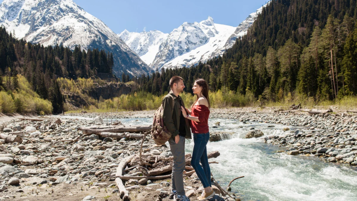 A couple stands on a rocky riverbank surrounded by lush green trees