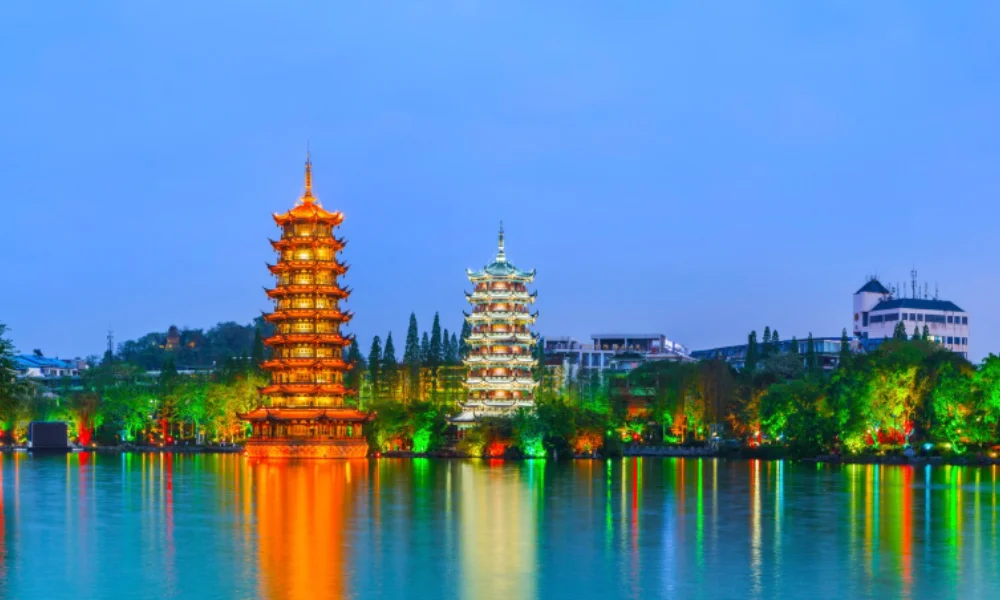 Illuminated twin pagodas at night reflect on a calm lake surrounded by vibrant greenery