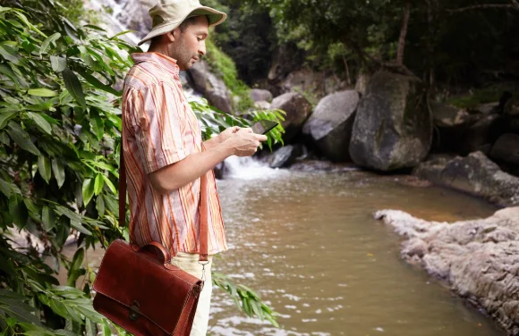 Man in a striped shirt and hat checks his phone by a peaceful river-south america travel destinations