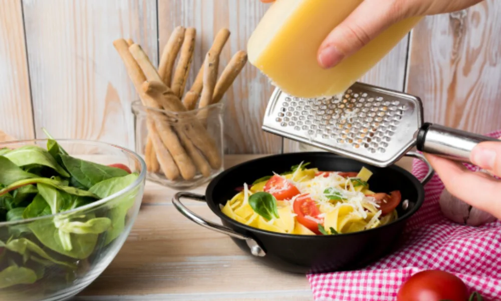 Hands grate cheese over a bowl of pasta with tomatoes and basil