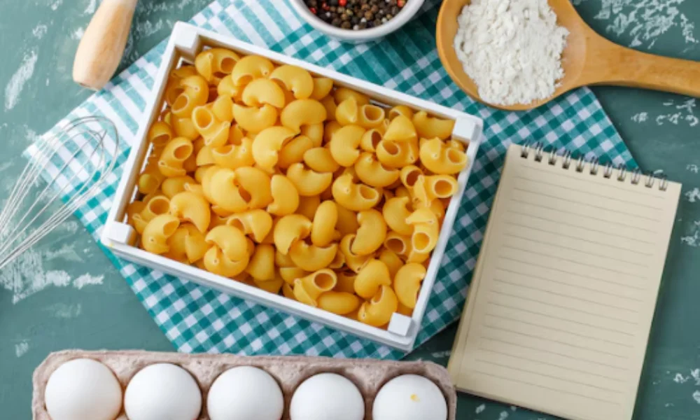 A box of uncooked macaroni sits on a checkered cloth surrounded by eggs