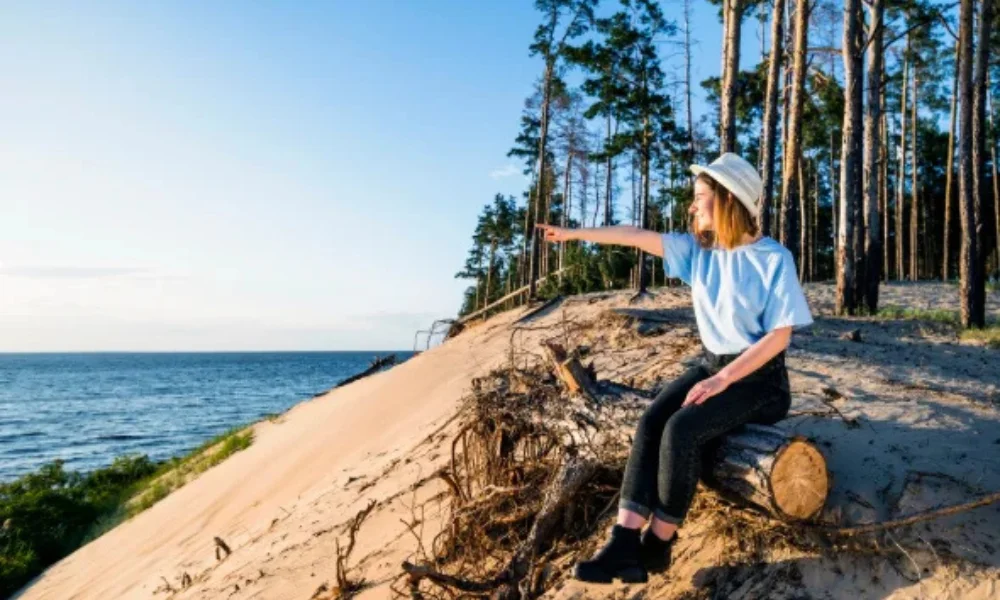 A woman in a hat sits on a log on a sandy beach
