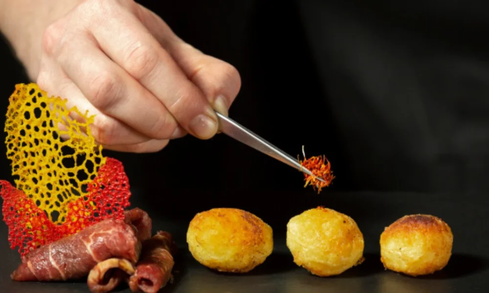 A chef's hand uses tweezers to place delicate garnish on three golden roasted potatoes beside artistic