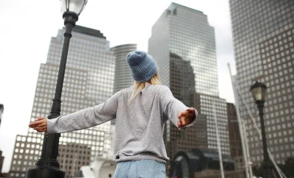 A person in a gray sweater and blue beanie stands with arms outstretched
