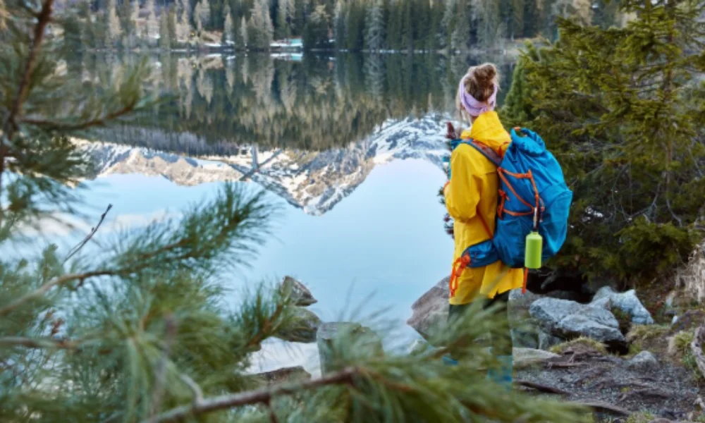 A hiker in a yellow jacket with a blue backpack stands by a calm lake reflecting snowy mountains