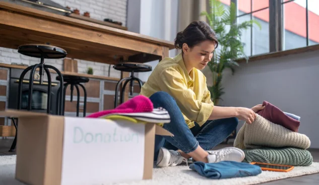 A woman in a yellow shirt sits on the floor, folding clothes next to an open donation box