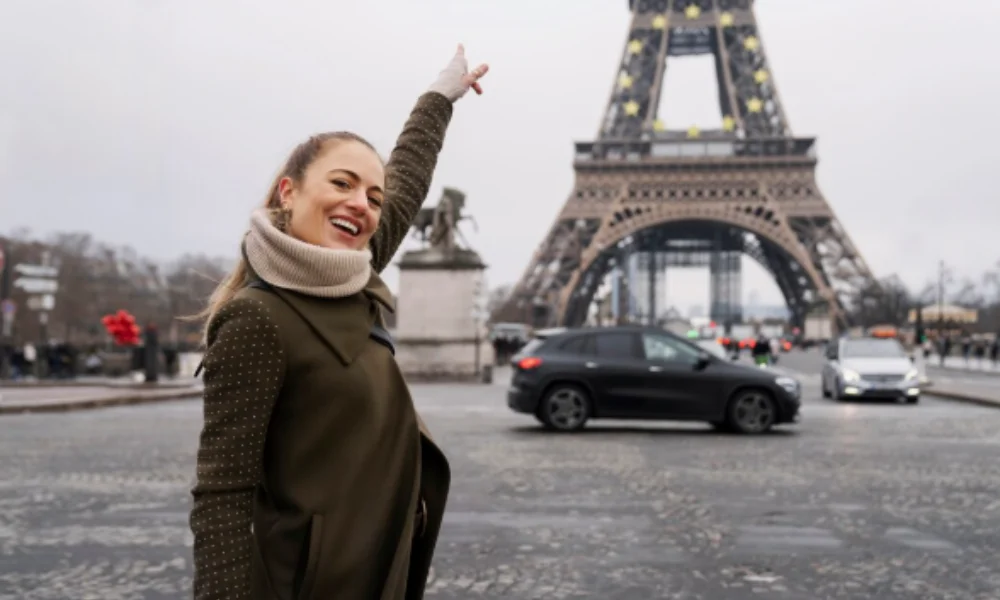 A smiling woman in a coat excitedly points up at the Eiffel Tower on a cloudy day in Paris-places to visit in france