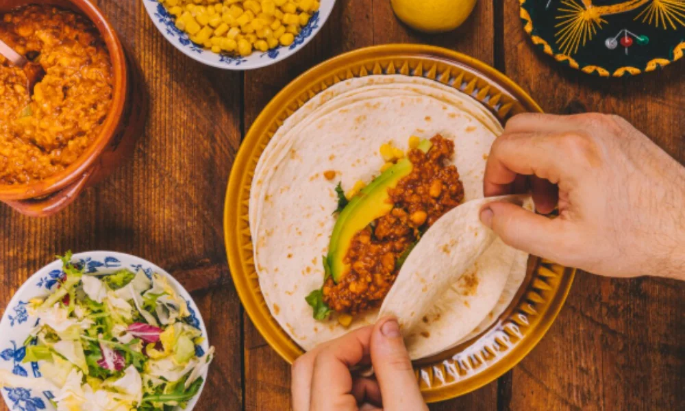 Hands roll a tortilla with lentils and avocado on a yellow plate