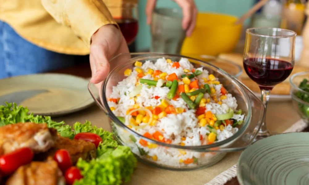 A hand places a bowl of vibrant rice salad with vegetables on a dining table, surrounded by chicken-easy chicken fried rice recipe