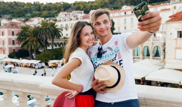 A smiling couple takes a selfie on a sunny balcony overlooking a charming Mediterranean town with colorful buildings