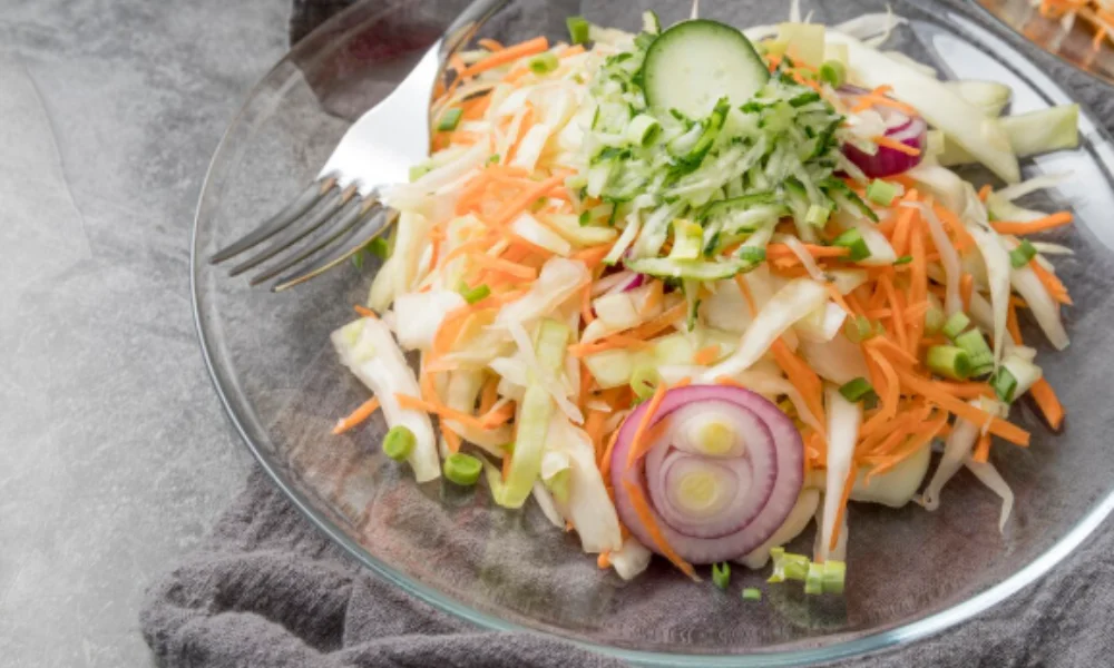A fresh salad on a clear glass plate features shredded cabbage, carrots, sliced cucumber, and red onion