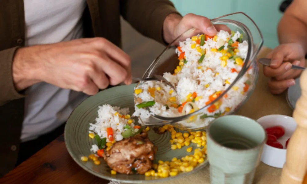 A person serves rice with mixed vegetables and corn onto a green plate with chicken