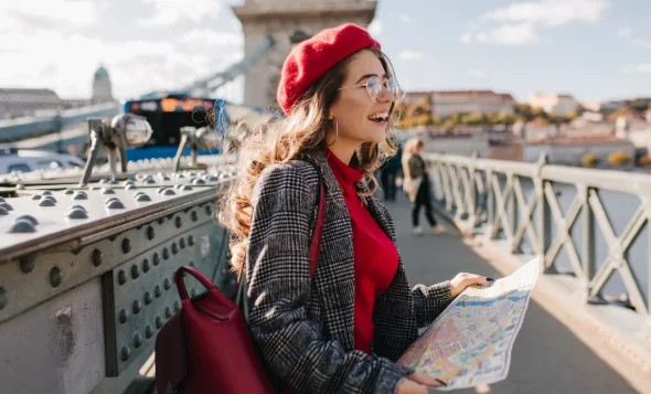 A woman in a red beret and glasses joyfully holds a map on a bridge, dressed stylishly in a plaid coat-travel destinations in europe