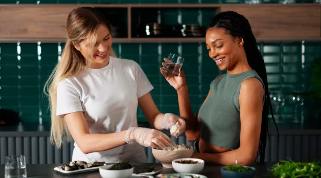 Two women in a kitchen, one wears gloves preparing food, smiling-travelling around the world