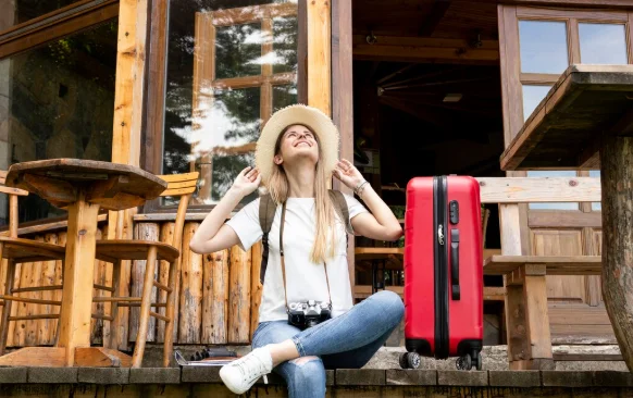 A woman in casual attire, wearing a sunhat, sits joyfully on wooden steps outside a rustic cabin-bucket list travel destinations