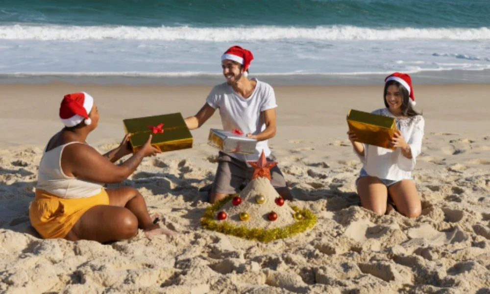 Three people wearing Santa hats exchange gifts on a sunny beach