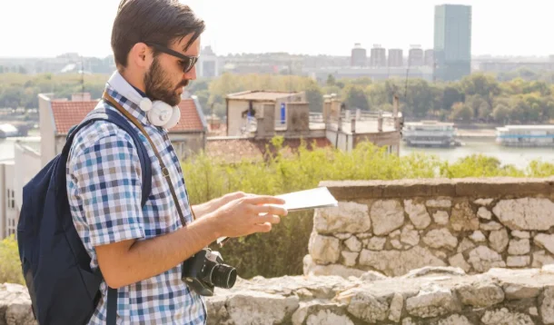 A bearded man wearing sunglasses and headphones stands outdoors holding a tablet-cheap travel destinations in Europe