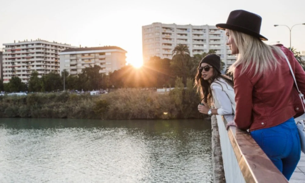 Two women stand on a bridge, gazing over a calm river at sunset